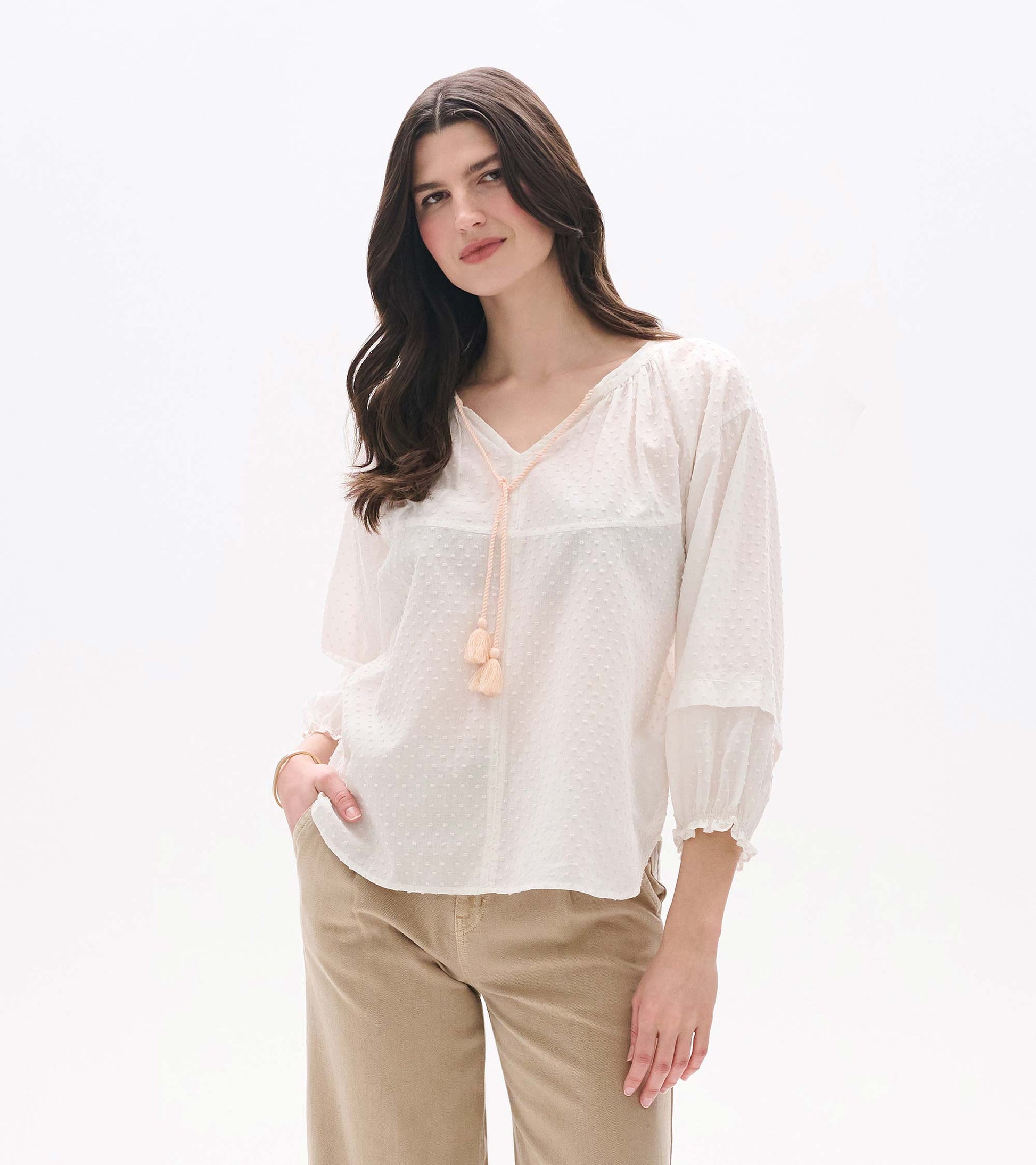 Blush Swiss dot blouse with V-neck, tassel tie, and long puff sleeves, worn with beige pants.