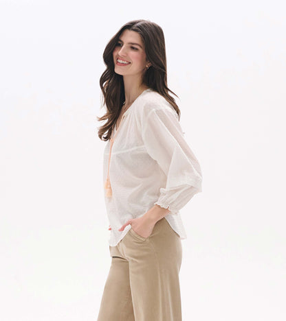 Woman wearing a blush Swiss dot blouse with sheer sleeves, V-neck tassel tie, and relaxed fit, side view