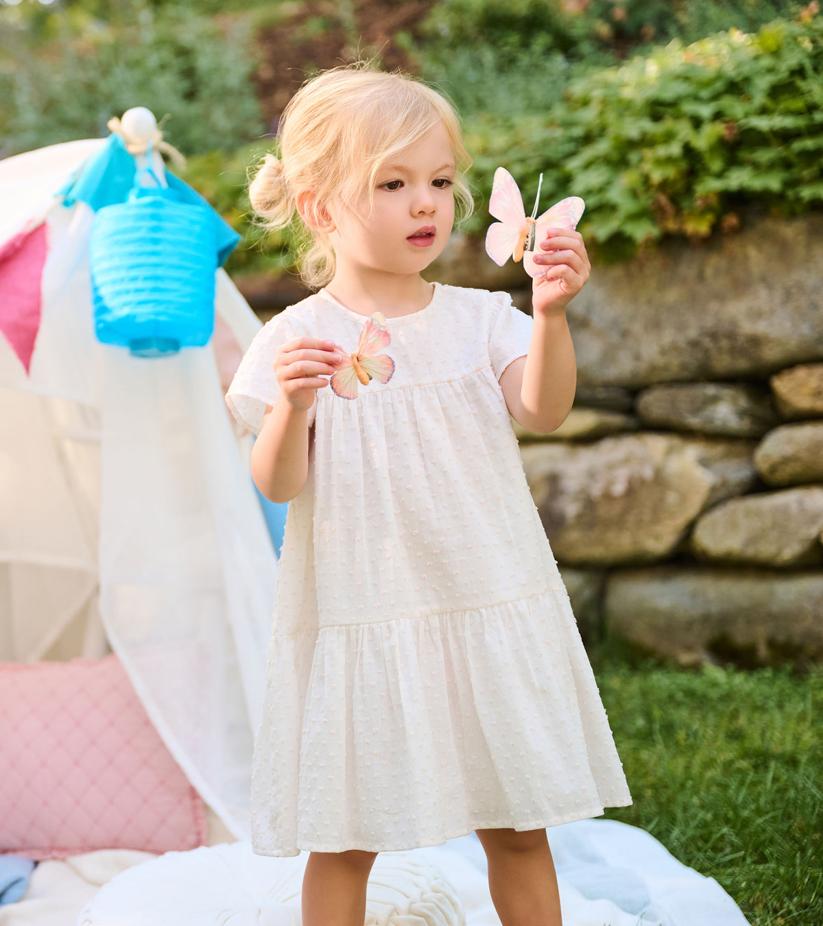 Toddler in an ivory Swiss dot dress holding butterfly toys, showing the front and tiered skirt