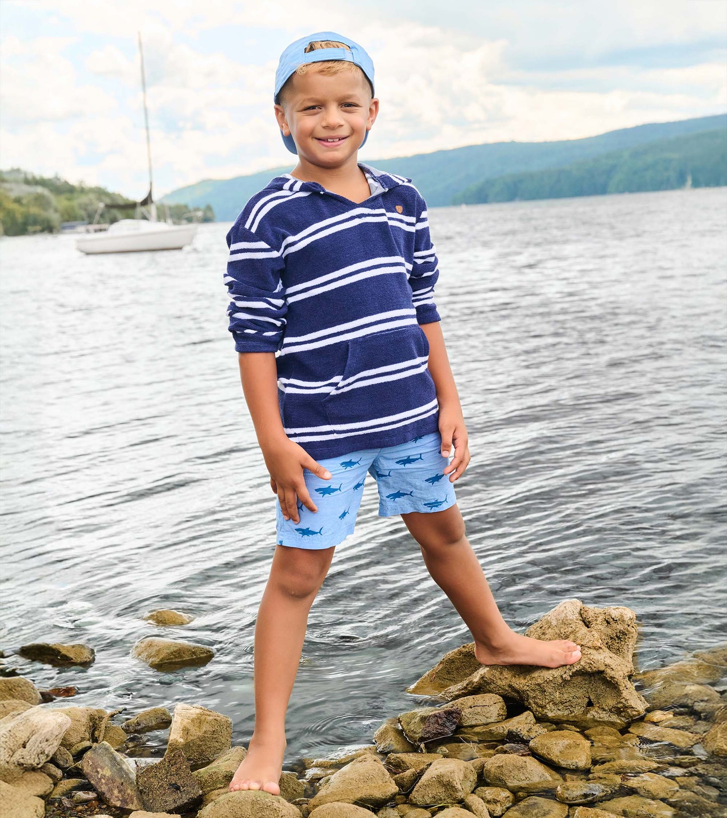 Boy wearing a navy and white striped hooded long-sleeve cover-up with front pocket, standing on rocks by the water.