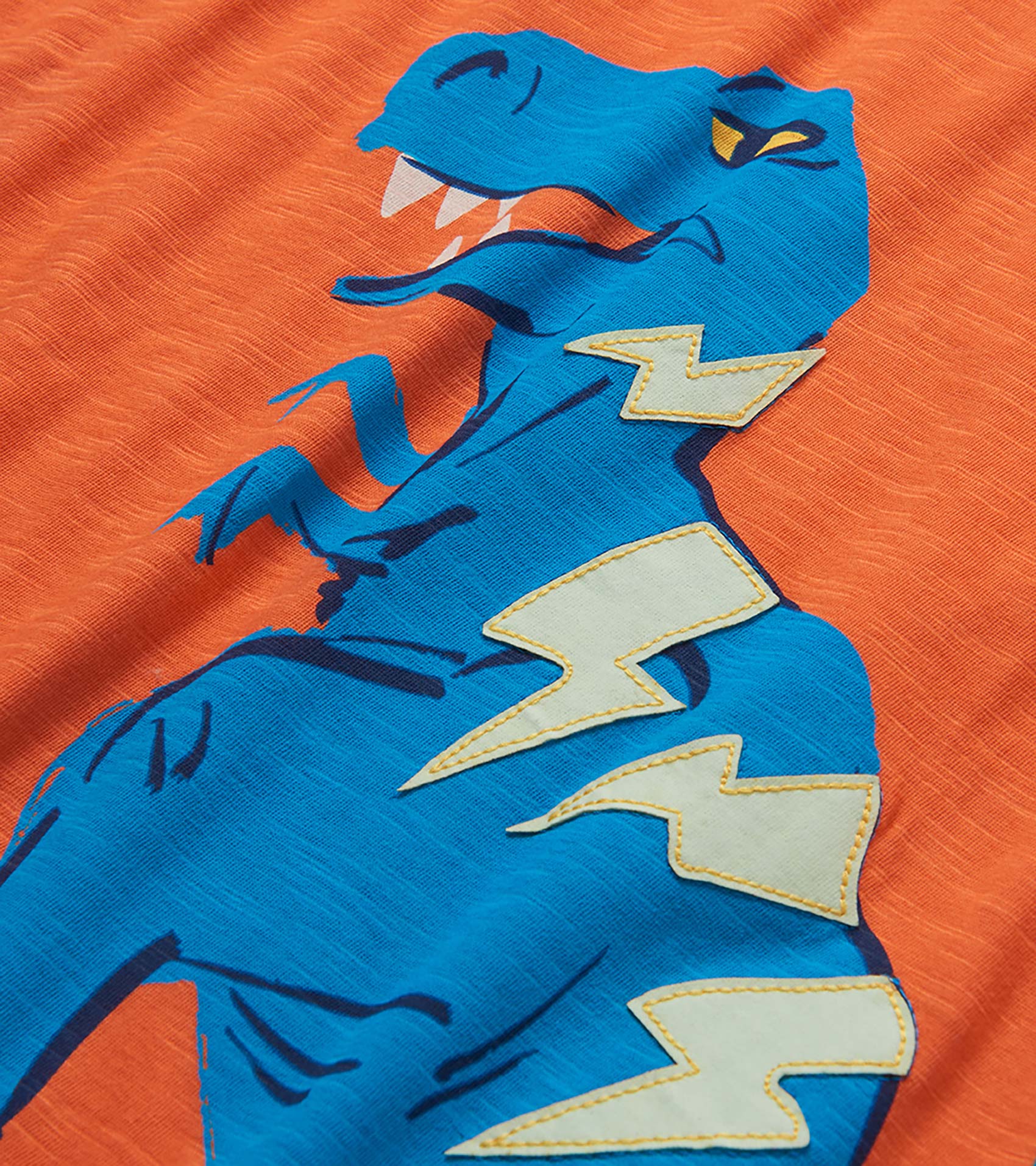 Blue dinosaur graphic on orange fabric with white lightning bolts, close-up of a T-shirt print