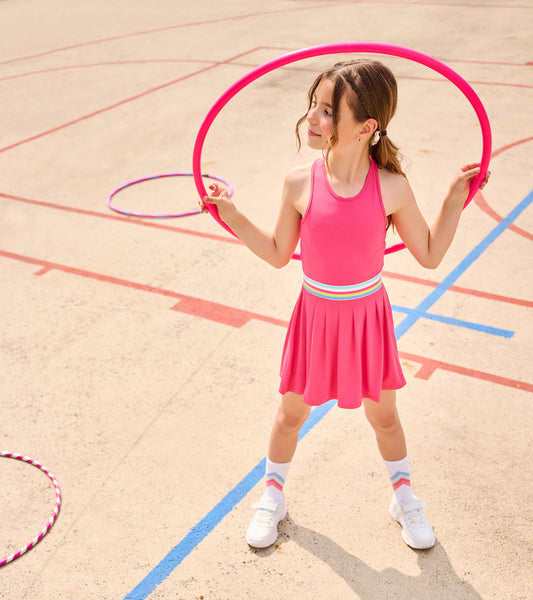 Girl wearing a bright pink sleeveless pleated active dress with a multicolor striped waistband