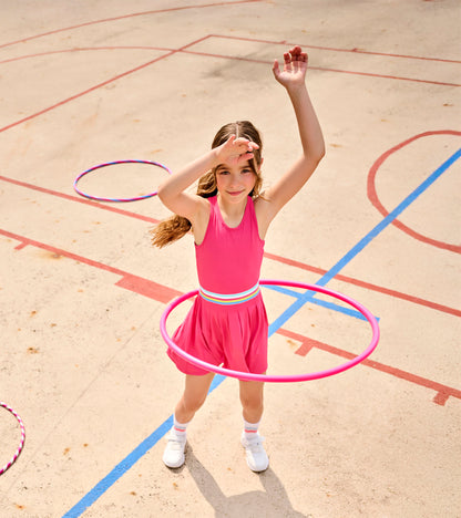 Girl wearing bright pink active dress with pleated skirt and striped waistband, hula hoop at waist