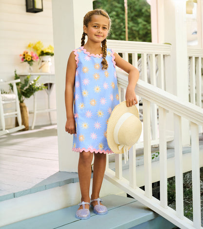 Girl wearing a light blue pinafore dress with pink trim and pastel sun print, holding a straw hat