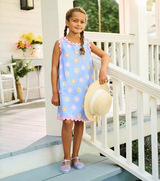 Girl wearing a light blue pinafore dress with pink trim and pastel sun print, holding a straw hat