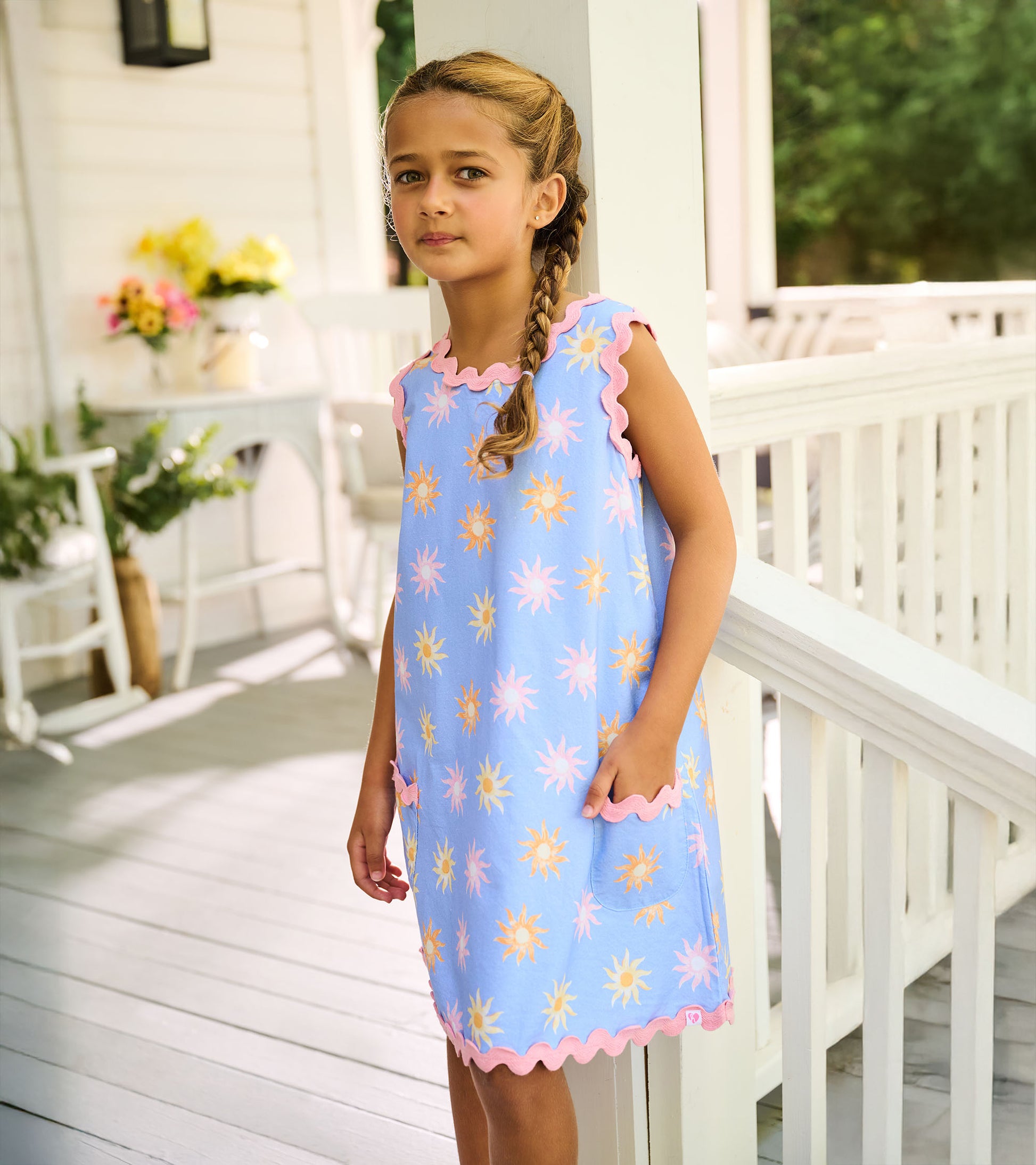 Side view of light blue pinafore dress with pastel sun print, pink scalloped trim, and patch pocket