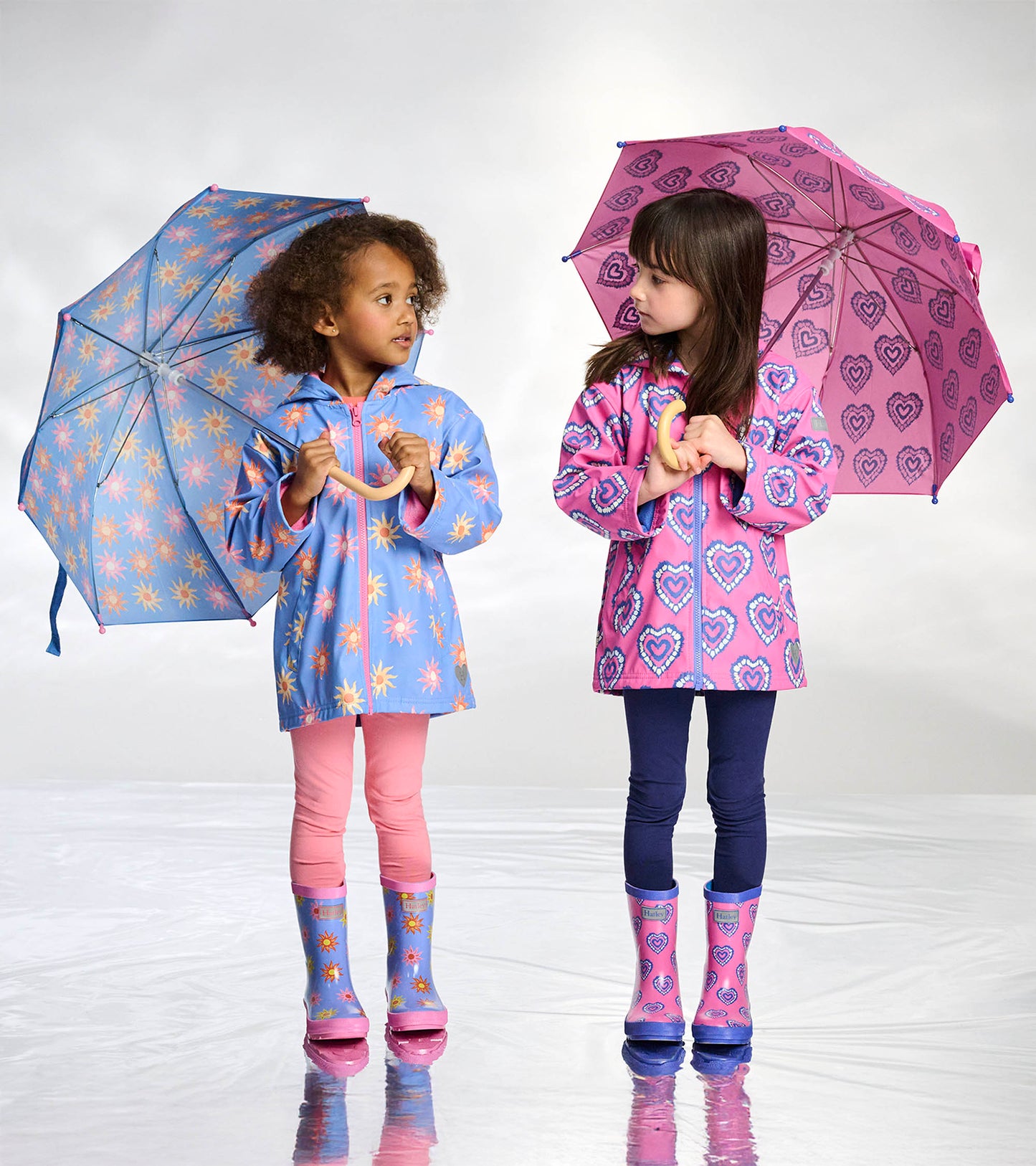 Two girls holding open umbrellas, one blue with sunburst print and one pink with heart print, wearing raincoats and boots