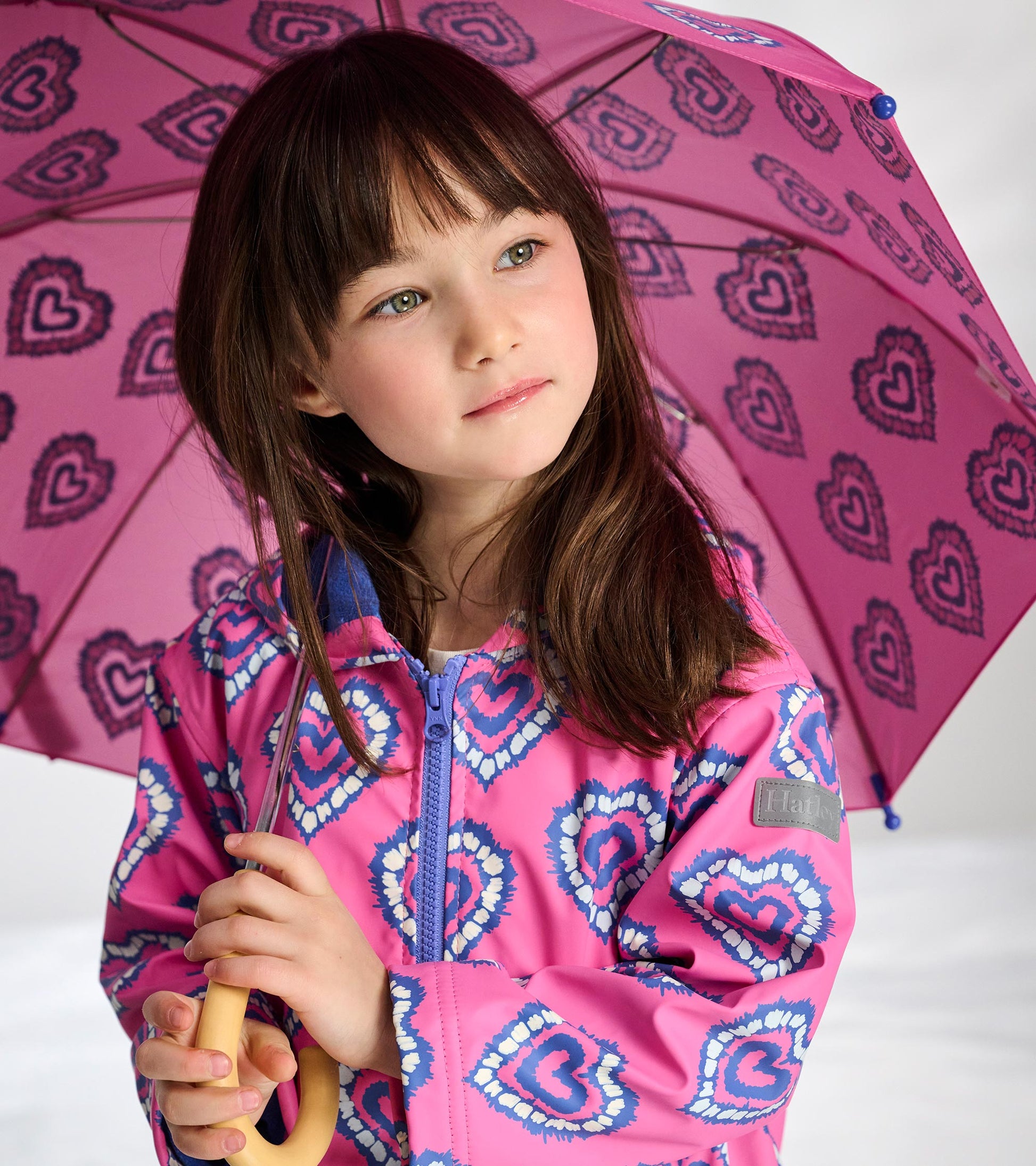 Girl wearing a pink heart-pattern zip-up rain jacket holding a matching pink umbrella, front view