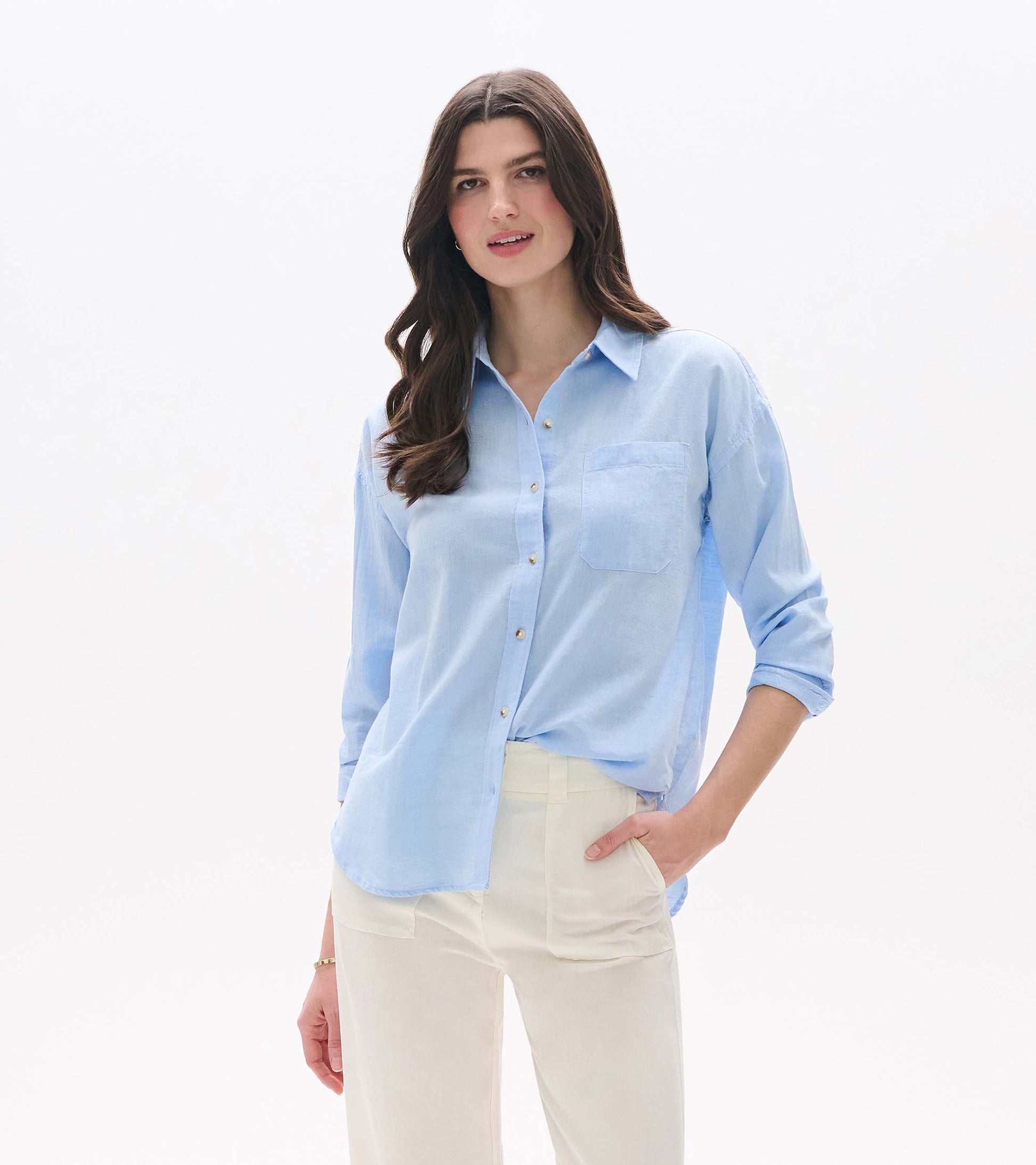 Light blue chambray button-down shirt with long sleeves, chest pocket, and relaxed fit, worn with cream pants.
