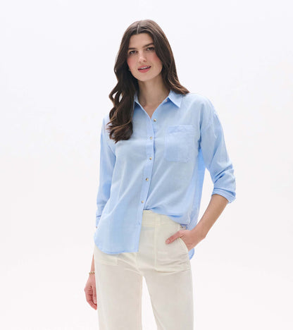 Light blue chambray button-down shirt with long sleeves, chest pocket, and relaxed fit, worn with cream pants.