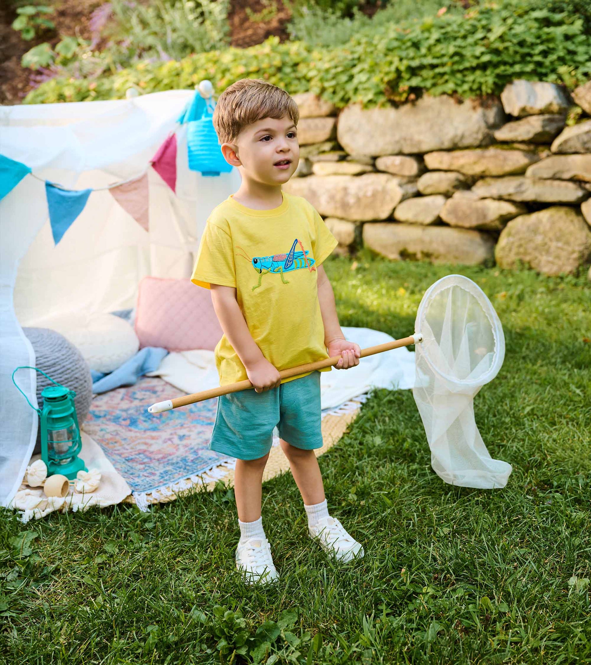 Toddler boy wearing a yellow grasshopper T-shirt and light blue shorts, holding a bug net outdoors