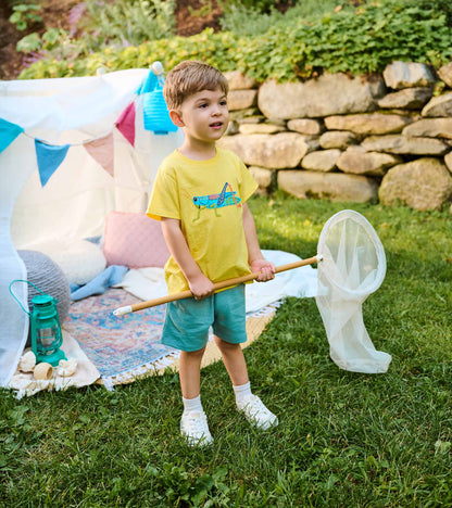 Toddler boy wearing a yellow grasshopper T-shirt and light blue shorts, holding a bug net outdoors