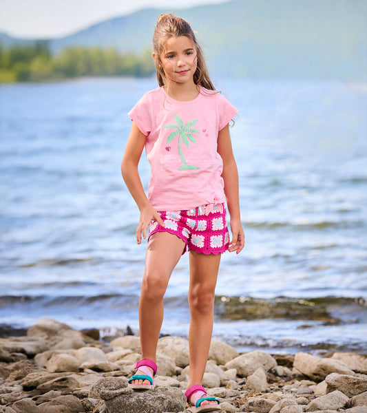 Girl wearing a pink short sleeve tie-front T-shirt with a palm tree graphic, standing by a rocky lakeshore