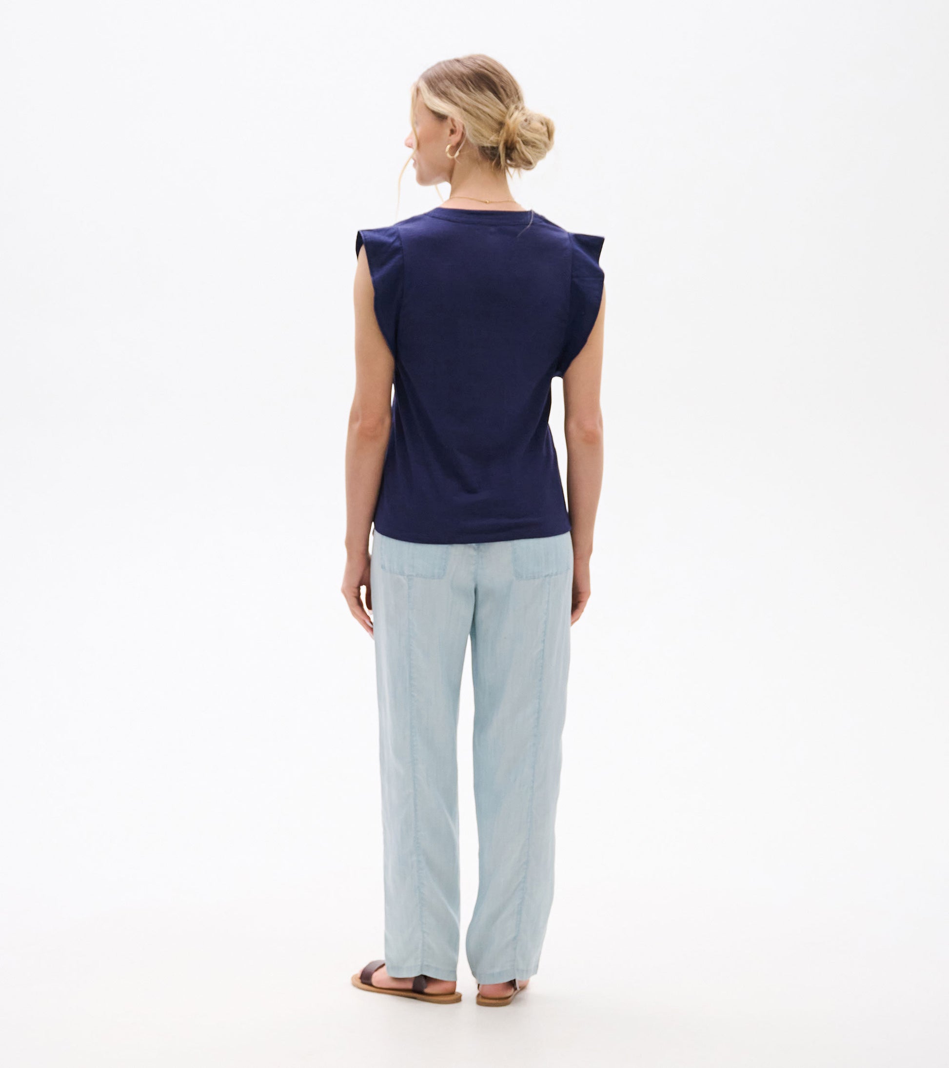 Back view of a navy sleeveless tank top with ruffled shoulder details, worn with light blue pants.