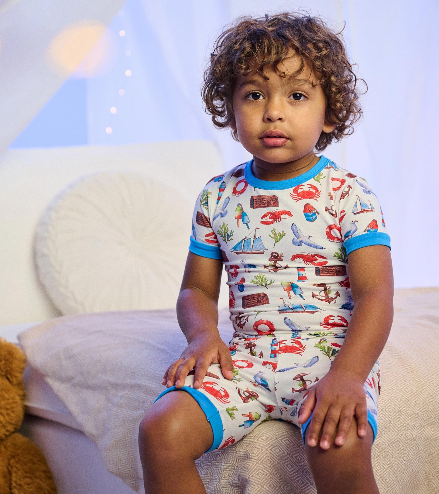 Boy wearing white short pajama set with colorful seascape print and bright blue trim, seated on a bed