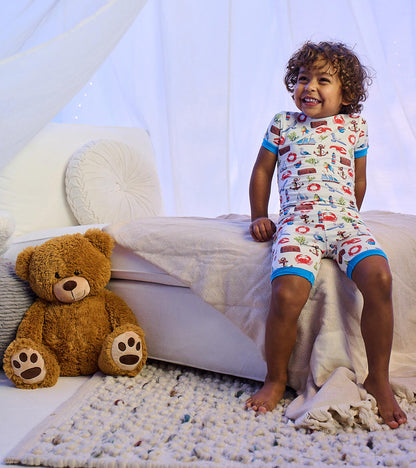 Toddler wearing white seascape-print short pajama set with blue trim, sitting on a bed