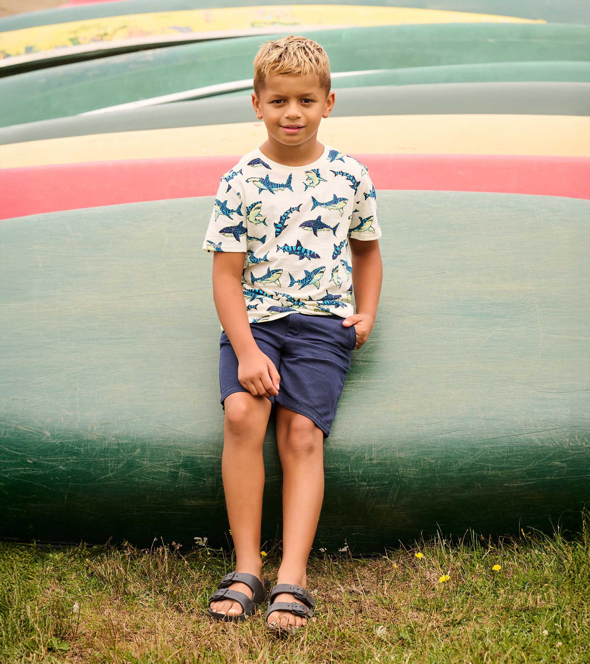 Boy wearing a cream short sleeve tee with blue shark print, shown outdoors with navy shorts