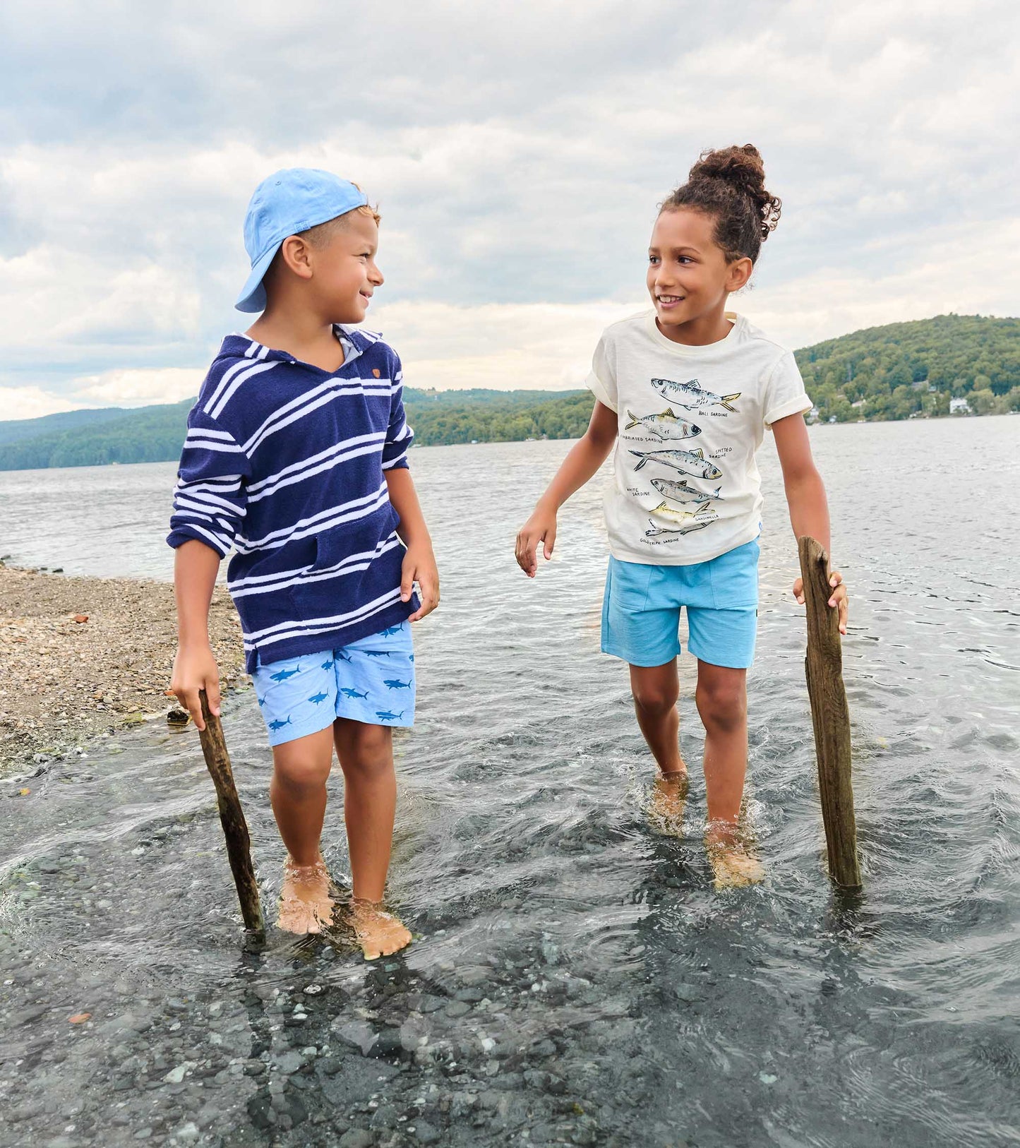 Two boys wearing graphic tees and shorts standing in shallow water by a lake