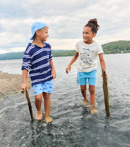Two boys wearing graphic tees and shorts standing in shallow water by a lake