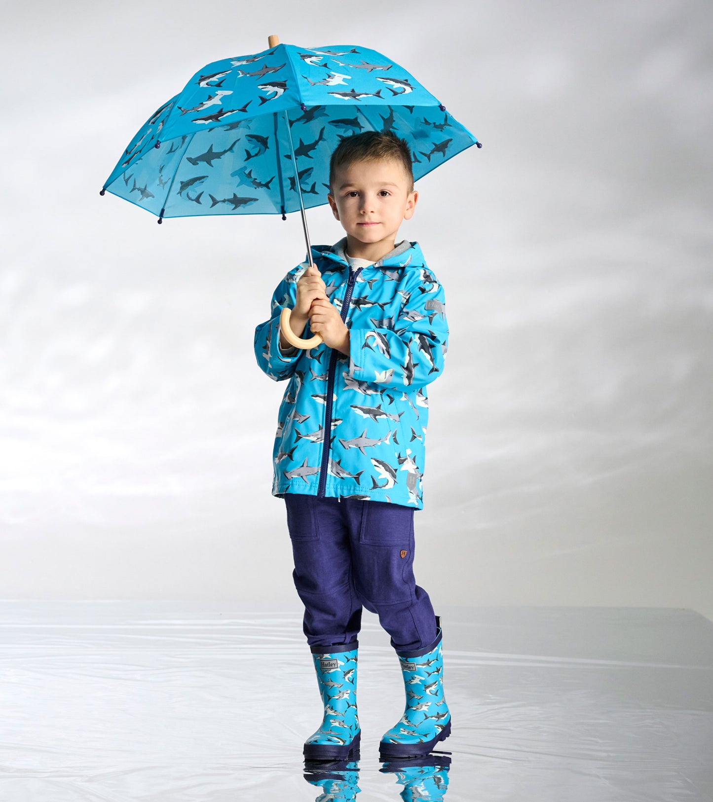 Boy holding a bright blue umbrella with shark print, wearing matching shark raincoat and boots