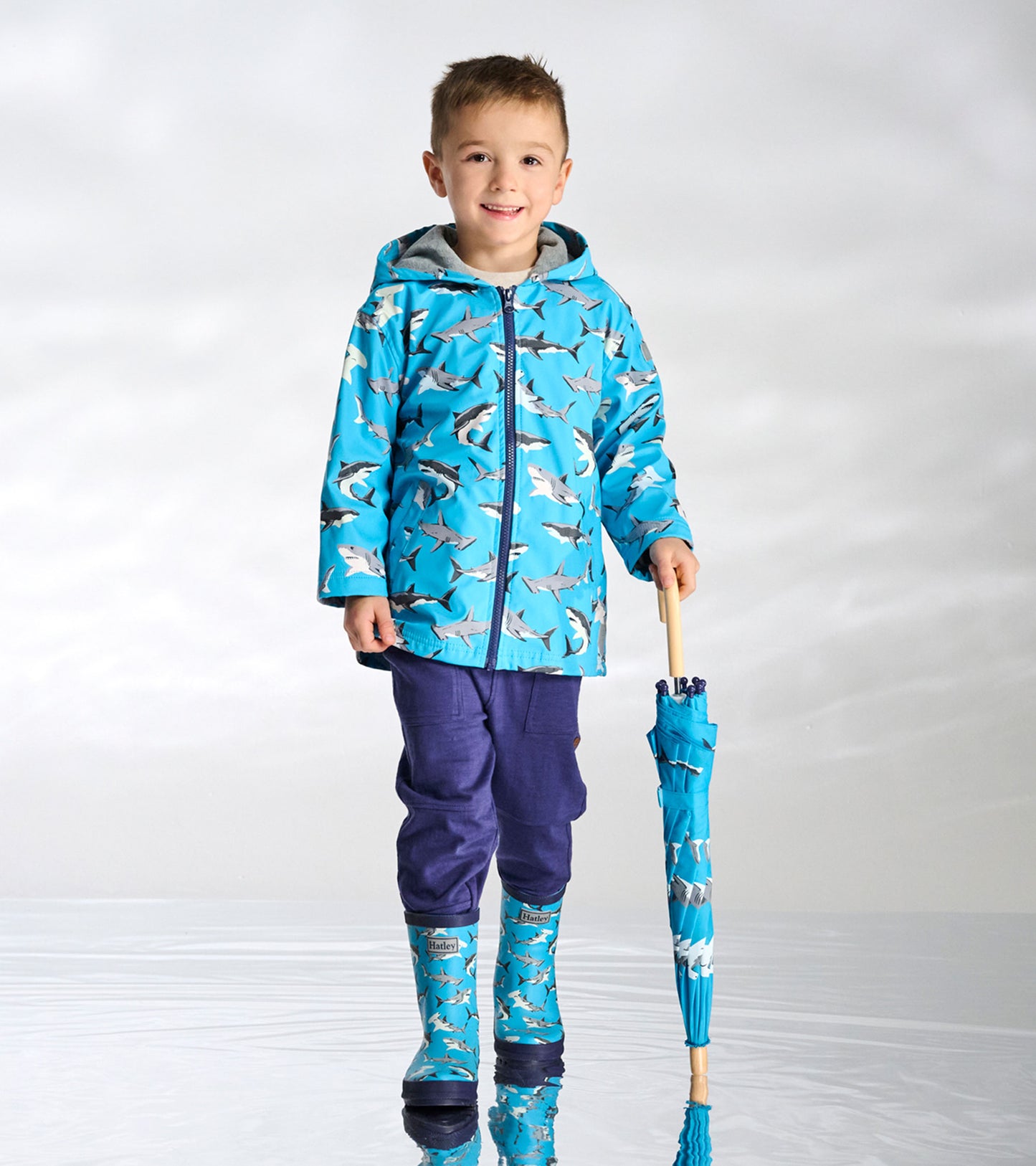 Boy wearing a blue shark-print zip-up rain jacket with hood, shown front view, holding a matching umbrella.