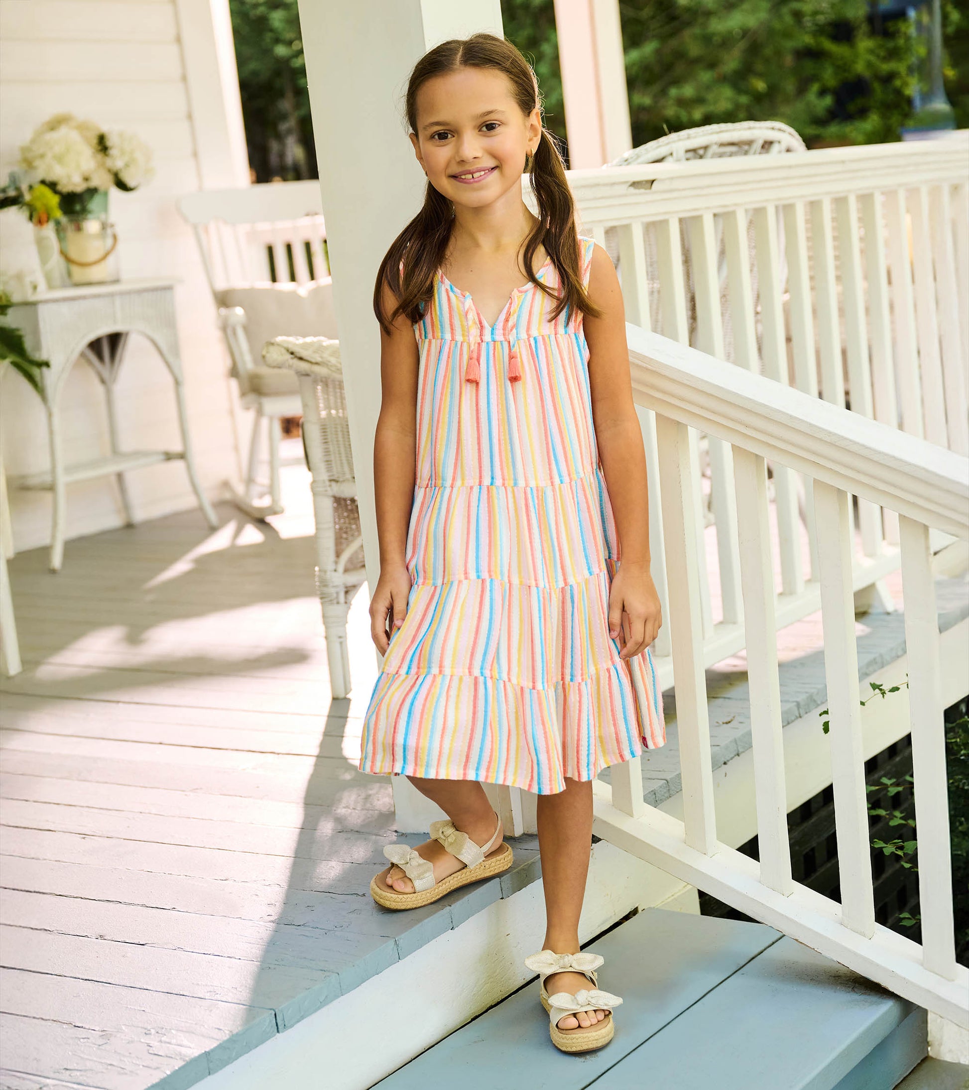 Girl wearing a sleeveless tiered dress with pastel vertical stripes and tassel ties at the neckline
