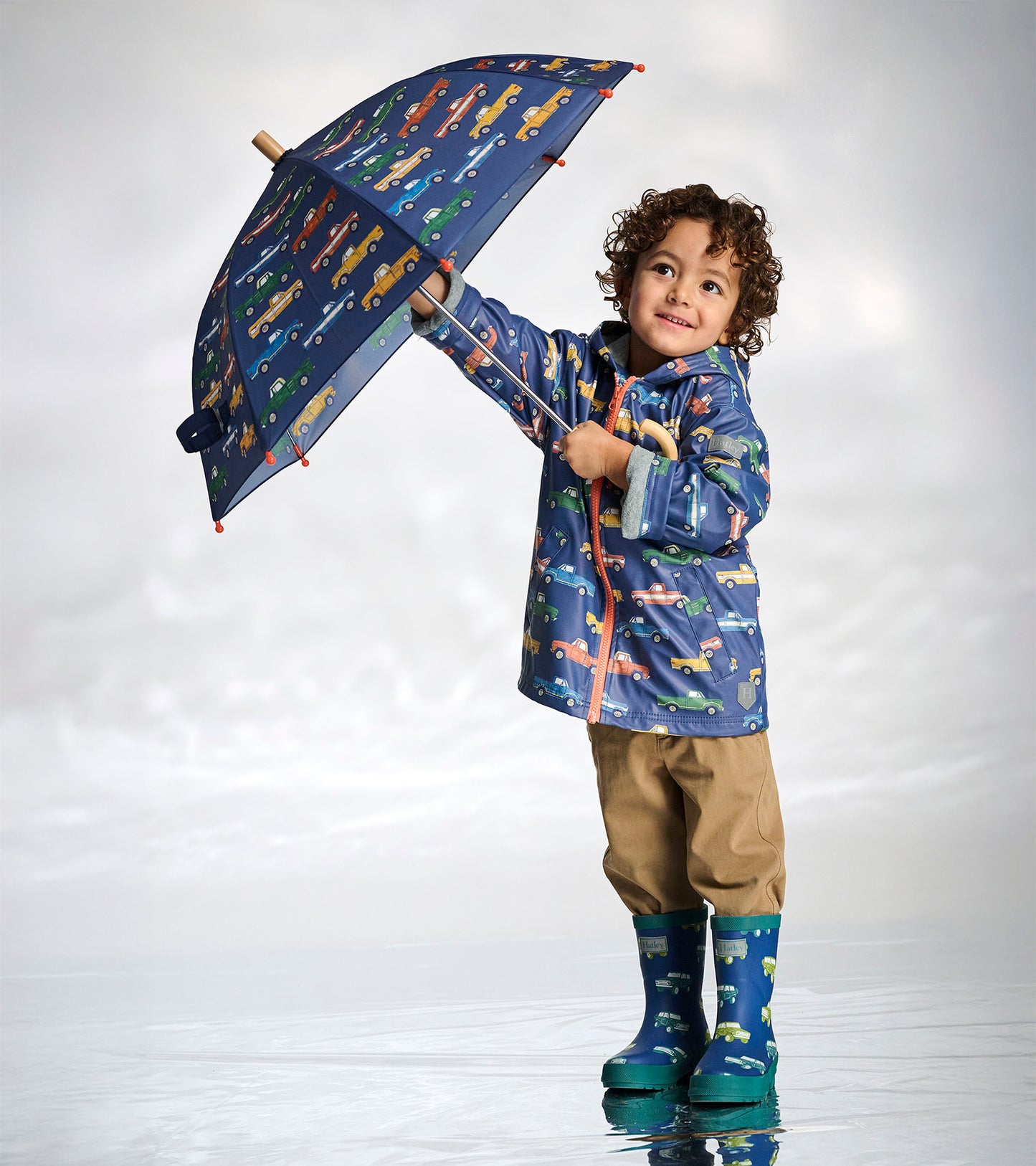 Child holding a navy umbrella with colorful truck pattern, wearing matching rain jacket and boots