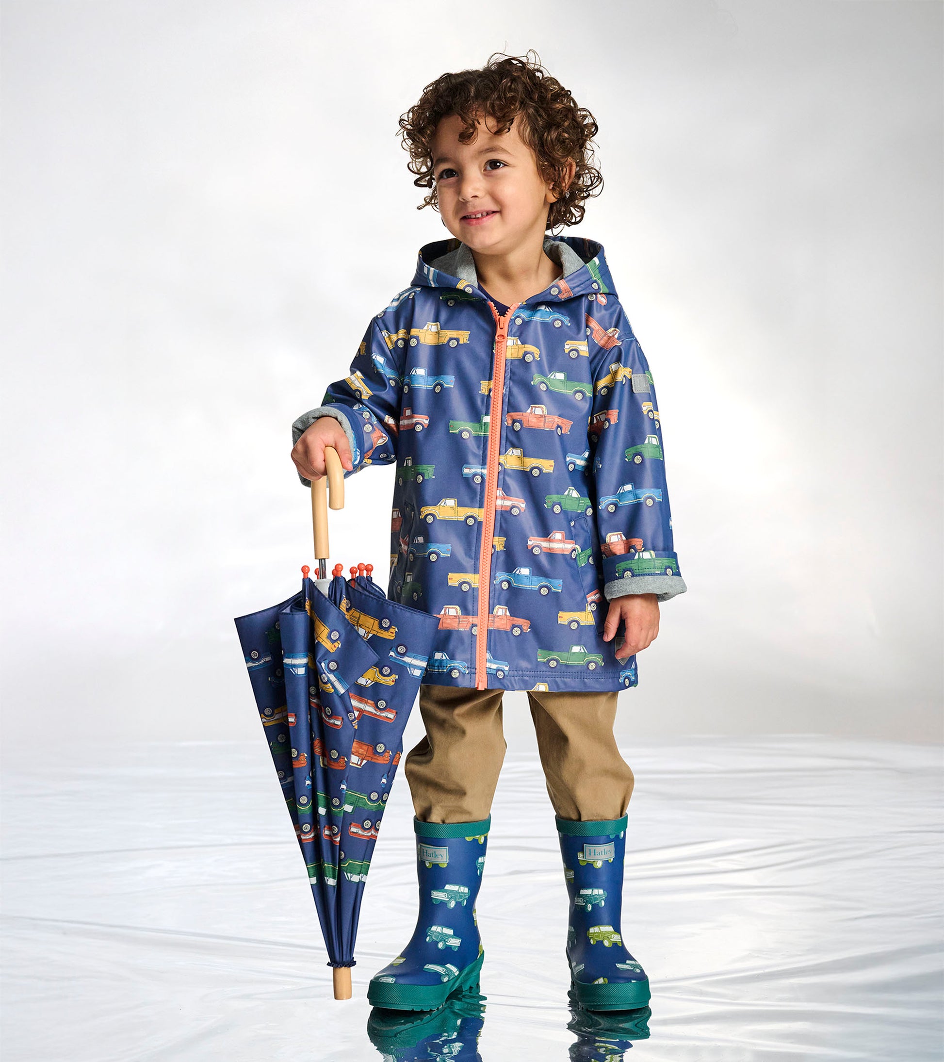 Blue umbrella with colorful truck print and wooden handle, shown folded and held by a child in rain boots