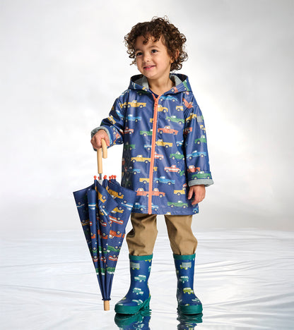 Blue umbrella with colorful truck print and wooden handle, shown folded and held by a child in rain boots