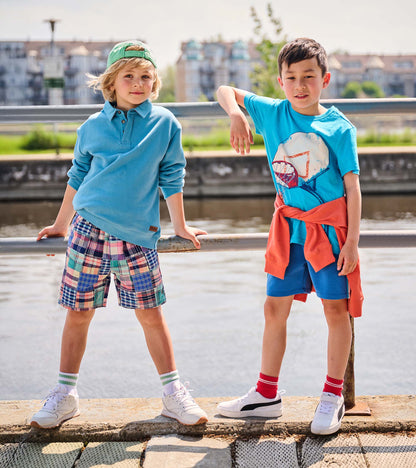 Boys wearing colorful plaid woven shorts, shown on a child standing outdoors by the water.