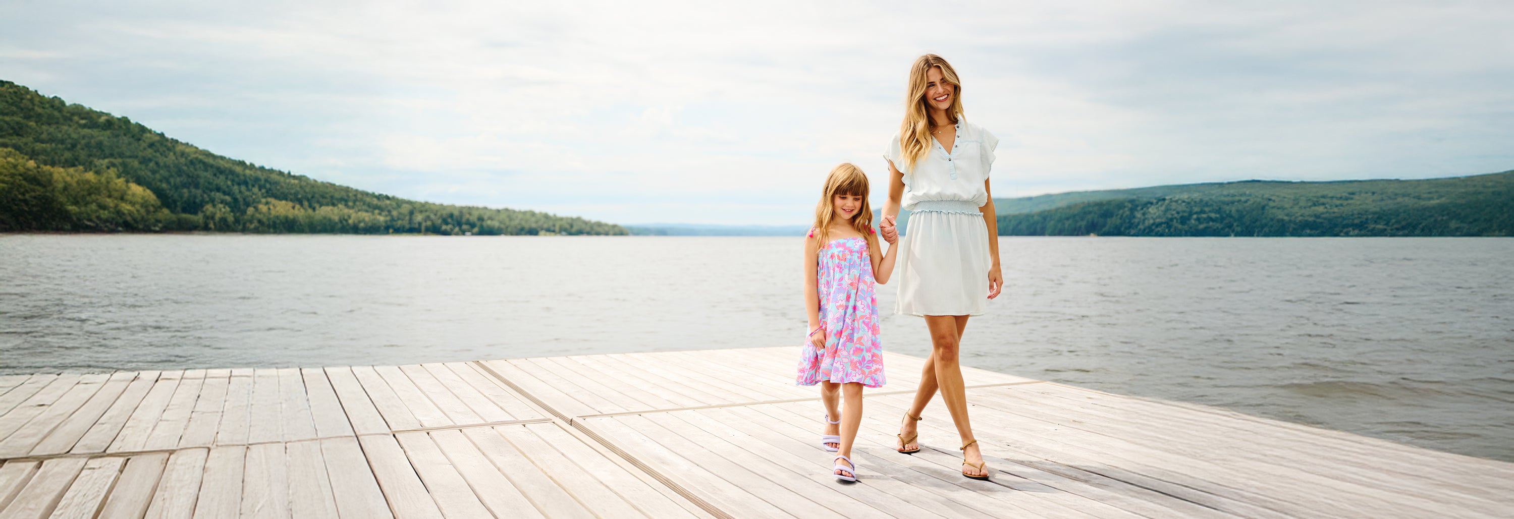 Promotes a relaxed family fashion collection. Showing woman and girl walking hand in hand on a lakeside dock.