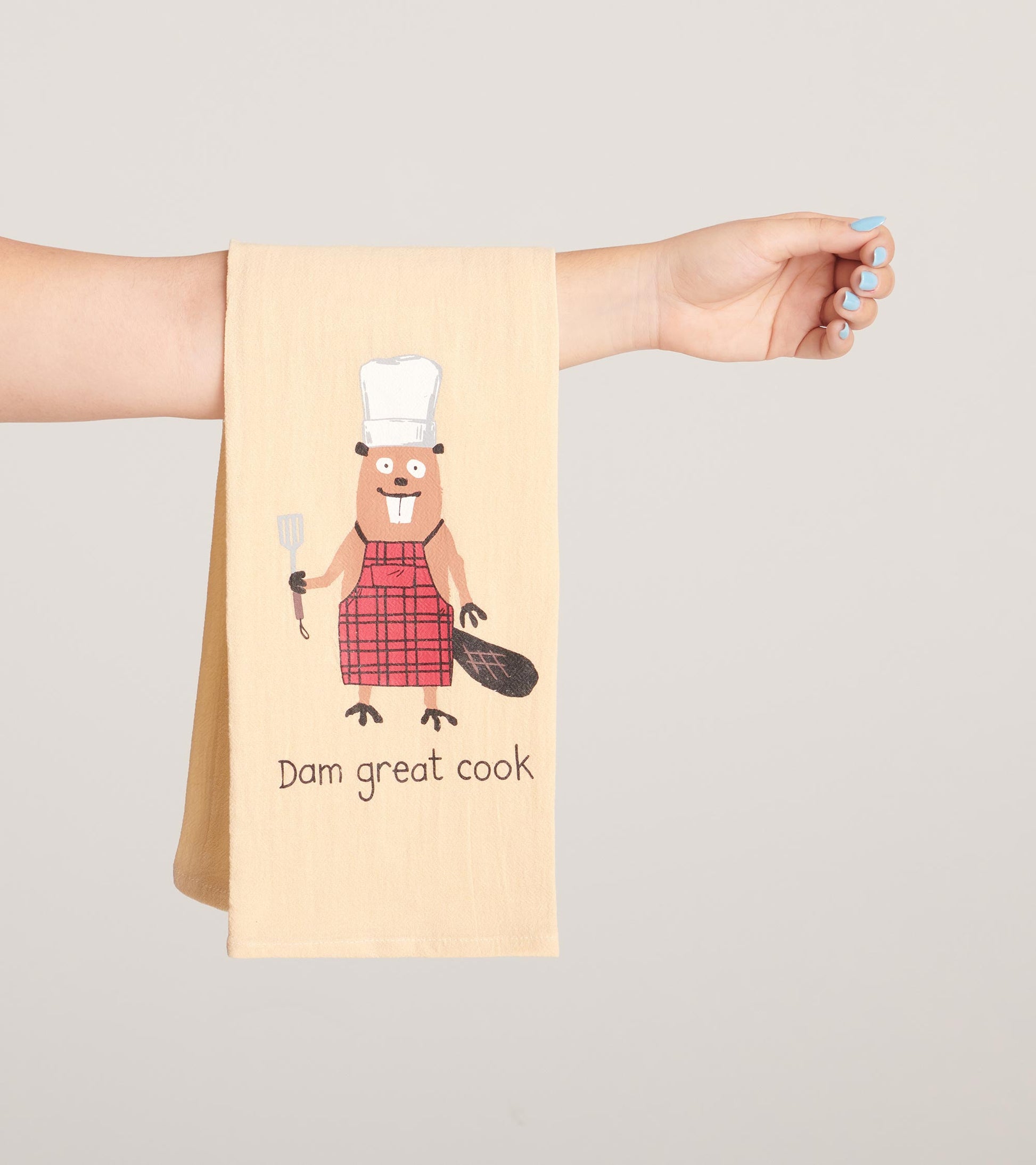 Beige tea towel featuring a cartoon beaver in a red plaid apron, holding a spatula with the text "Dam great cook.