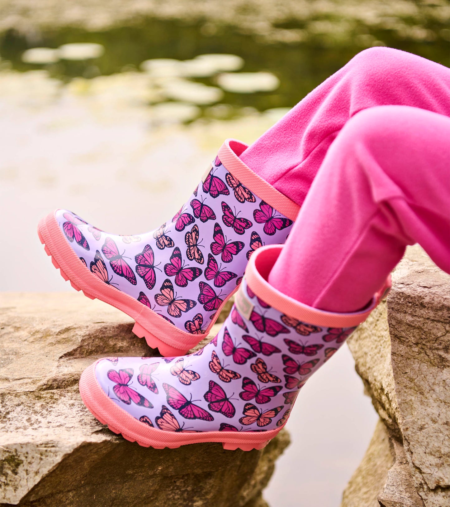 Lavender toddler rain boots with pink butterfly print and pink trim, shown on a child’s feet outdoors