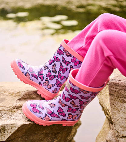Lavender toddler rain boots with pink butterfly print and pink trim, shown on a child’s feet outdoors