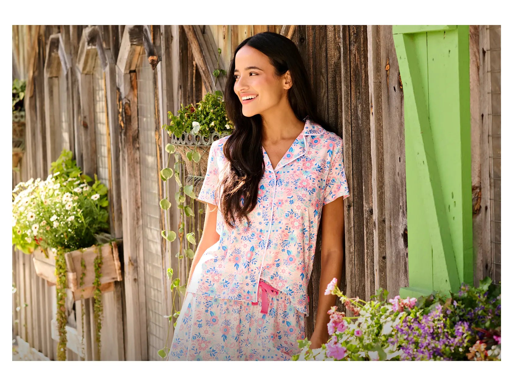 Promotes pajama sets. Showing woman in a pink floral pajama set standing by a rustic wall with flowers.