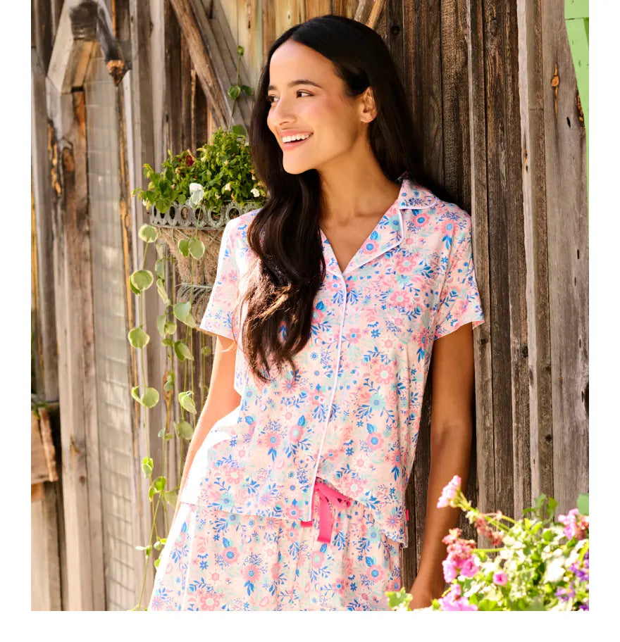 Promotes women's pajama sets. Showing woman wearing a pink floral short-sleeve pajama set by a rustic wall.
